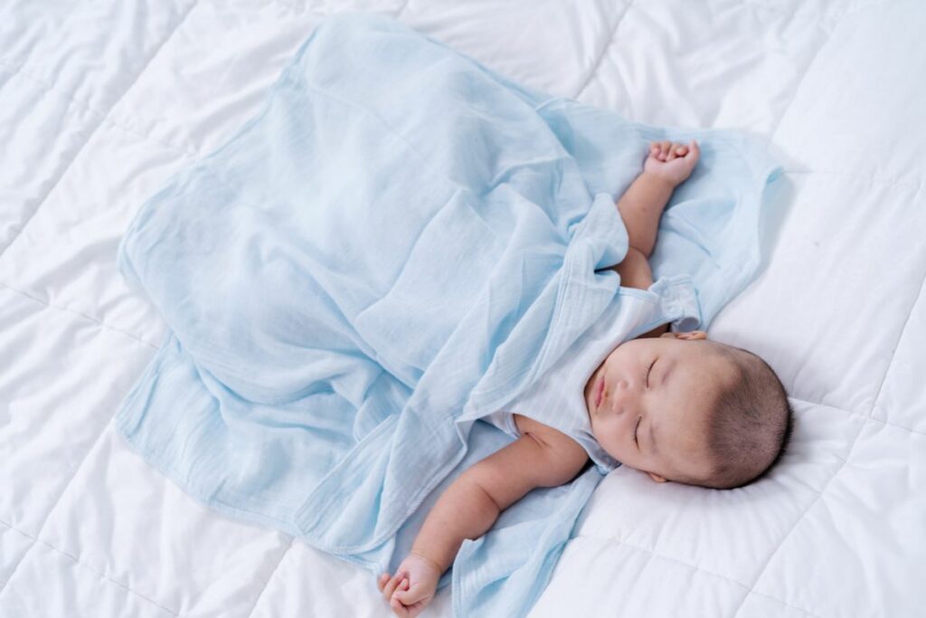 A baby peacefully sleeping in a blue blanket on a white bed mattress